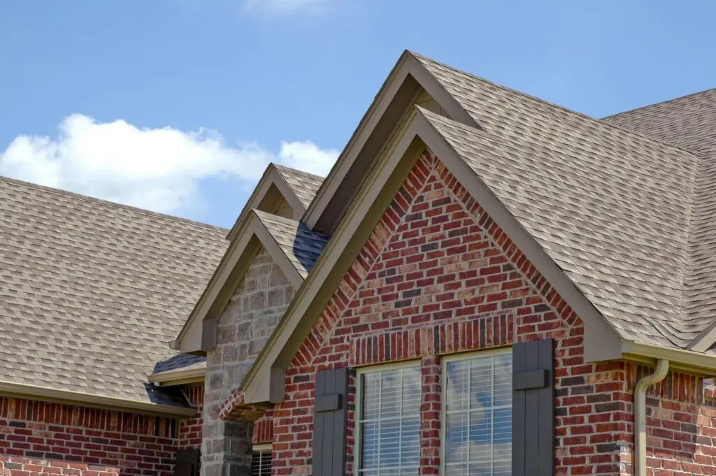 Seamless beige gutters along a brick home's roofline, with layered gable peaks and asphalt shingle roofing