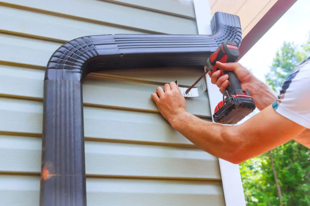 Technician using a drill to secure a downspout bracket during gutter installation on vinyl siding