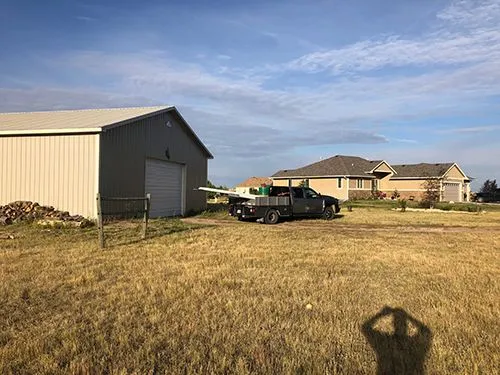 Metal pole barn with a work truck parked beside it, with a nearby home visible across the field
